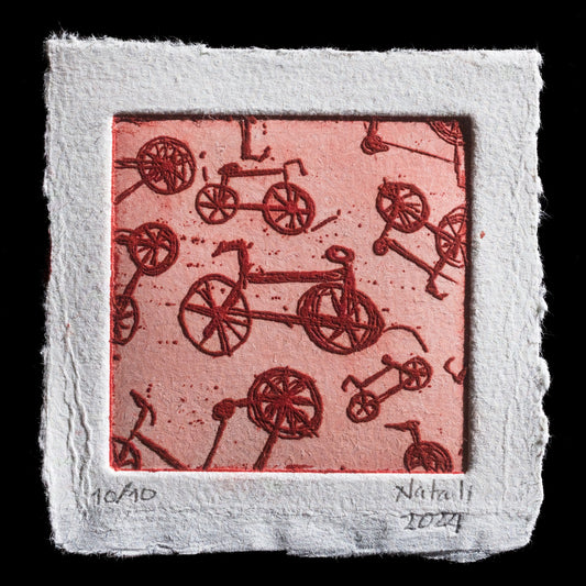 This etching by Natali Osorio Estrada showcases a repeating pattern of bicycles on a pink background, surrounded by an off-white border. The bicycle wheels are depicted as circles with spokes, while the frames are simplified to emphasize movement through bold lines and minimal details. The overall effect is one of dynamic energy, capturing the essence of cycling in motion.