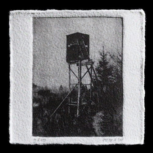 The print 'On the lookout' by Monique van de Wijdeven showcases a dark forest scene with a deer stand in the center, surrounded by trees. The overall mood of the artwork is somber and contemplative. Created using intaglio and photopolymer techniques, this piece presents a dramatic black and white landscape that invites viewers to reflect on their surroundings.