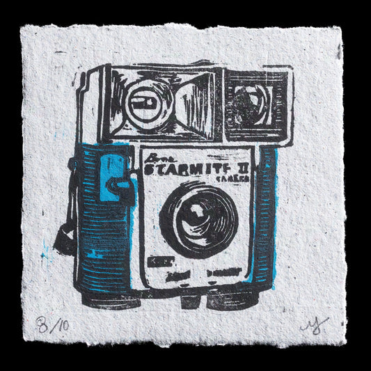 The artwork 'Starmite' is a linocut print by Melissa Jaarmaasa. It features a vintage camera as its central motif against a light grey background. The black ink adds depth to this monochromatic piece, highlighting the intricate details of the camera's design.