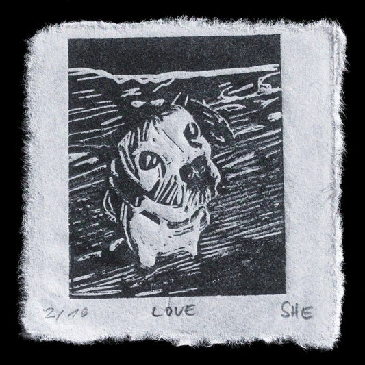 This black-and-white relief print, "Love" by "she", features a stylized head with an open mouth and closed eyes, set against a dark background. The subject's face is rendered in bold lines, while their hair flows outward from the top of their head. The print is signed 'SHE' at the bottom right corner, accompanied by handwritten notes that read '21/20' and 'LOVE'.