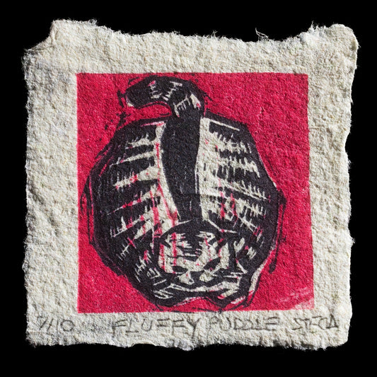 This linocut print, "Fluffy Puddle" by Juliana Steca, features a whimsical depiction of a puddle with soft, rounded edges and a subtle gradient effect achieved through the use of red ink on textured paper. The image is framed by a white border, adding to its playful character.