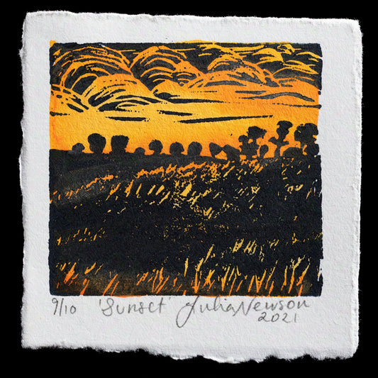 This linocut print by Julia Newson captures a serene sunset scene, characterized by warm hues and gentle, wispy clouds in shades of yellow and orange. The image features silhouettes of trees and hills in black, set against a gradient sky that transitions from light to dark. The artist's signature and edition number are discreetly placed at the bottom edge, providing context without detracting from the natural beauty of the print.