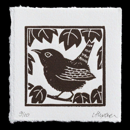 This linocut print, "Little Wren" by Alexandra Owen, showcases a small bird amidst foliage in rich dark brown hues against a crisp white background. The artist's signature is discreetly placed at the bottom right corner.