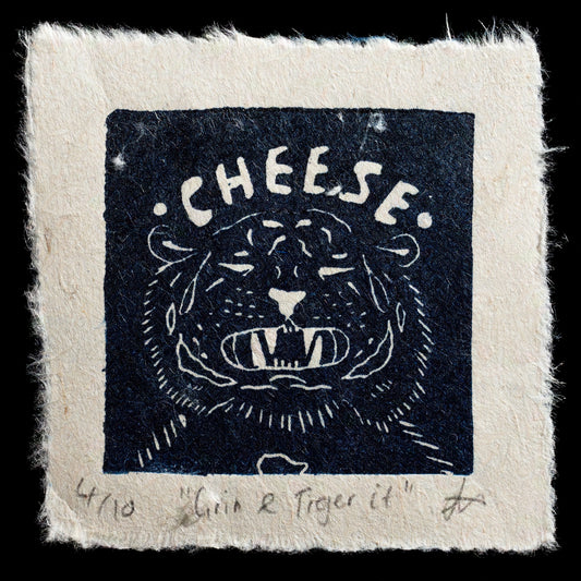 This captivating print by Jennifer Mottram is a linocut titled "Grin and Tiger it". The image features a tiger's head with its mouth open in a cheerful grin, set against a black background. The word "CHEESE" arches above the tiger's head, adding to the playful atmosphere.