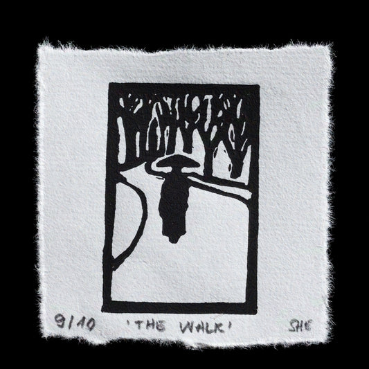 This linocut print, titled "The Walk" by artist "she", features a serene black-and-white composition. The design depicts two figures strolling through a forest, surrounded by trees and foliage. A subtle border frames the artwork, adding depth to its overall aesthetic.