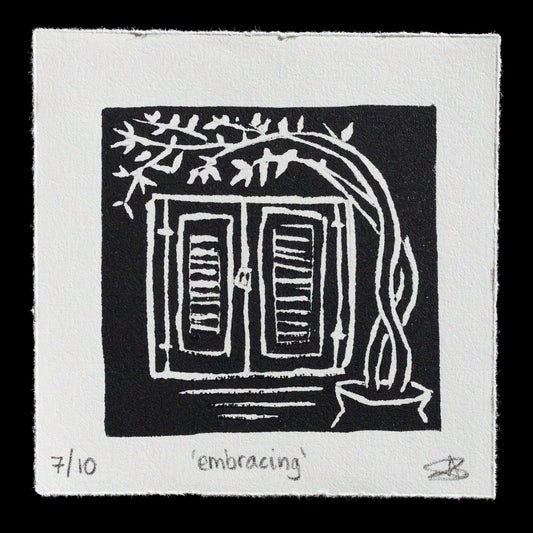 This print, titled "Embracing" by Antigoni Vlachou, features a striking black-and-white linocut technique. The composition showcases a window with open shutters, surrounded by lush greenery, creating an inviting and serene atmosphere.