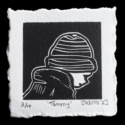 This linocut print, 'Tommy' by Susan Monaghan, features a monochromatic design in black ink on white paper. The artist has skillfully captured a figure wearing a striped beanie, showcasing their use of relief printing techniques to create intricate details and textures.