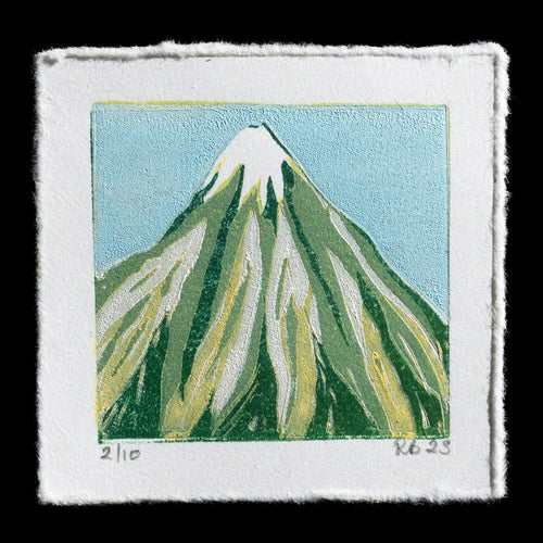 This linocut print depicts Mount Taranaki on a clear day, showcasing its snow-capped peak against a light blue sky with wispy white clouds. The mountain's slopes are rendered in shades of green and yellow, while the foreground features lush grasses and trees in various hues of green. A subtle shadow effect adds depth to the scene, which is set against a plain black background.