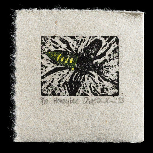 This linocut print, titled "Honey Bee" by Anita Fina Kiewra, showcases a detailed depiction of a honey bee in flight amidst foliage. The black linoleum print features subtle yellow accents on the wings and abdomen, while the background is a warm beige hue with an orange tone towards the bottom edge, adding depth to the composition. The square shape of the print measures 3 inches by 3 inches.