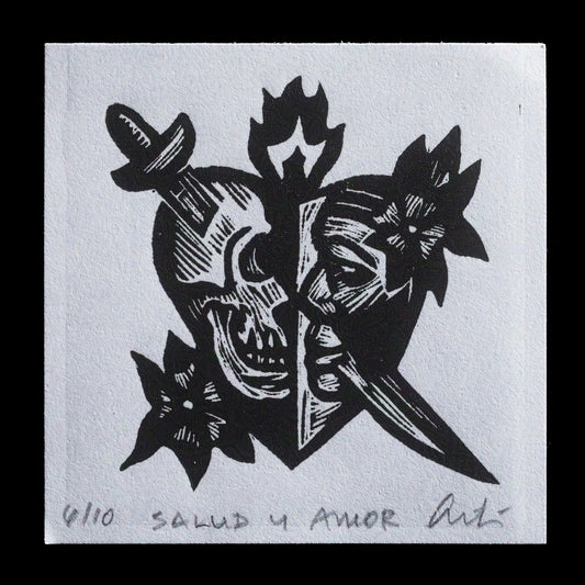 This linocut print by Adrian Tio, titled "Salud y Amor," showcases a striking heart-shaped design featuring two skulls facing each other in the center. The skulls are surrounded by flowers and leaves, with the phrase 'SALUD Y AMOR' inscribed at the bottom of the print in black ink on white paper.