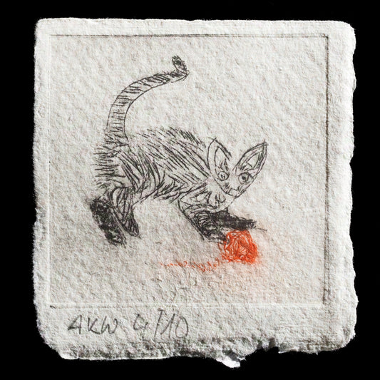 This etched print by Alain Wohlfarth depicts a young cat in play. The black-and-white image features a small orange ball at its feet, with subtle hints of red watercolor accents beneath the cat's paws. The artwork is presented on TetraPak paper.